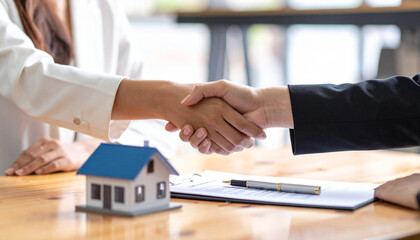 Real estate agent and client engaging in a handshake after finalizing a contract for a new home, with a miniature house and pen placed on the table, symbolizing a successful deal