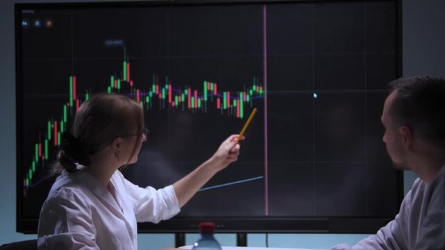Woman and man at table in high-tech office analyzing stock market charts on screen