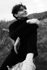 Fototapeta premium Emotion black and white portrait of a young guy in a black wear. Man have relax and smile. Seating on the tree