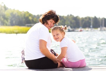 Obraz premium Mature woman and young girl sitting together on a dock by the water, enjoying a sunny day, sharing a moment of joy and connection with nature and family