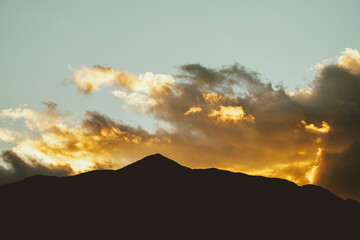 A silhouette of a mountain range is backlit by a golden sunset, with clouds adding texture to the sky.