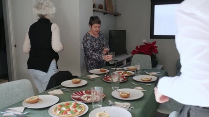 Multigenerational family gathering for festive winter holiday meal, carefully setting dining table with plates, glasses, bread, and traditional Christmas dinner preparations