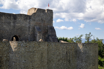 section of Neamț Fortress in Romania. Weathered stone walls and a tower with a lone flag reach towards a partly cloudy sky.