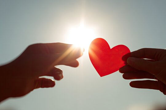 Two hands reaching for a red heart against a bright sky