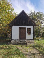  charming, small traditional Romanian house with white walls, a dark wooden roof, and a single window with flowers, set in a sunny autumn landscape.