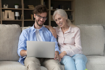 Modern tech time. Caring grownup son grandson sit on couch together with mature mum granny teach...