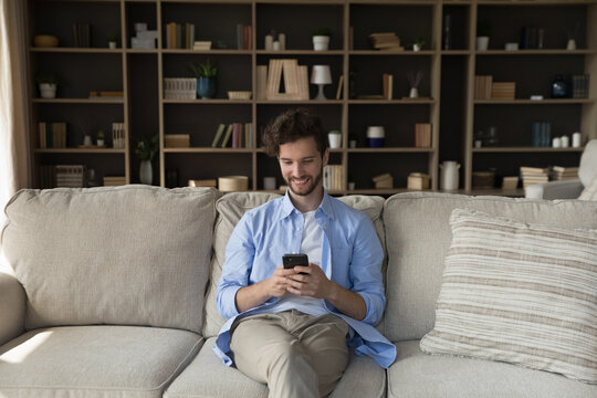 Comfy weekend with phone. Smiling young male hipster enjoy leisure time online use cellphone on comfy couch at home text message to girlfriend scroll ecommerce websites read news watch vlogs stories - Powered by Adobe