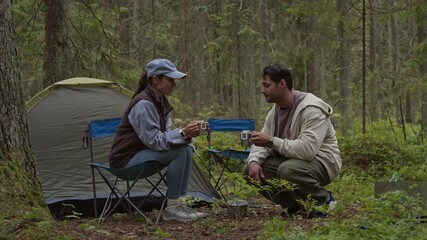 Side view of diverse young couple of travelers drinking drip coffee while having picnic by tent camping in green forest - Powered by Adobe