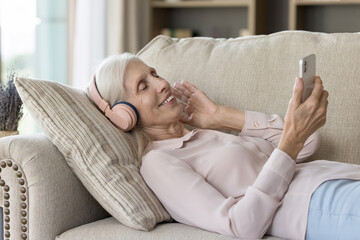 Connected leisure. Peaceful carefree older age female rest on comfy couch in digital earphones device choose favorite song from online player on mobile phone listen to calm melody enjoy soothing audio