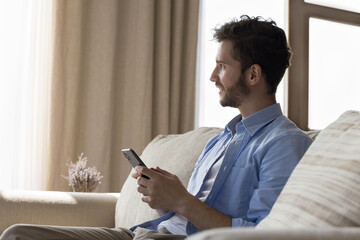 Tech connected lifestyle. Dreamy thoughtful gen z man rest on comfy sofa at home hold smartphone...