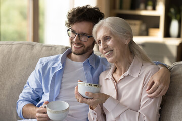 Warm vision of tomorrow. Affectionate young man grown child cuddle on couch with older adult mommy...