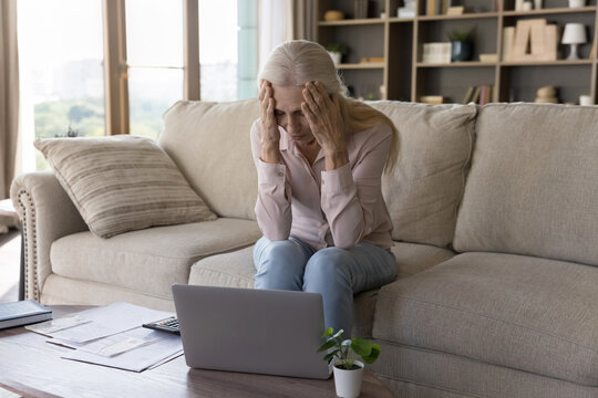 Financial stress. Frustrated depressed older woman retiree sit on couch in home office do paperwork hug head in anxiety overwhelmed by debt bankruptcy too high expenses lack of money on bank account - Powered by Adobe