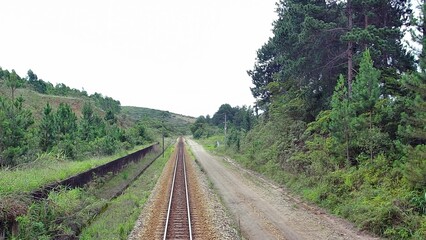 Obraz premium Expansive view of train tracks running through a rural landscape with lush green hills and a dirt road in Catas Altas, Minas Gerais, near Bicame de Pedra, Brazil. Overcast day, no people.
