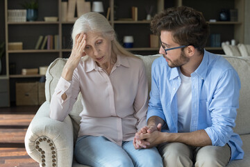 Tender family moment. Attentive compassionate grownup son sit on sofa with sad suffering senior mom...