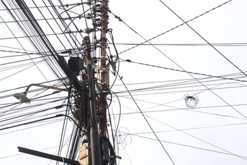 A chaotic cluster of tangled overhead wires and multiple rusty electric poles against a clear sky,...