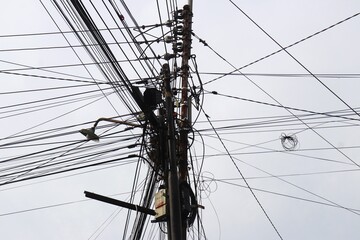 A chaotic cluster of tangled overhead wires and multiple rusty electric poles against a clear sky,...