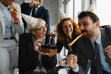 Group of business colleagues celebrating in a office with a birthday cake, party hats and blowers. The coworkers enjoying a fun in office party