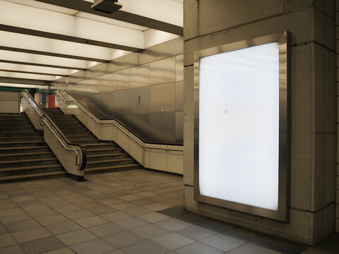 Blank Illuminated Vertical Billboard Mockup in Subway Station with Stairs, Metal Walls and City Transport Signage.