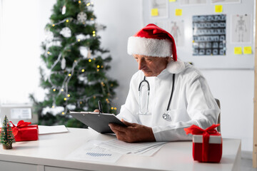 Mature male doctor in Santa hat reviewing medical record in his office