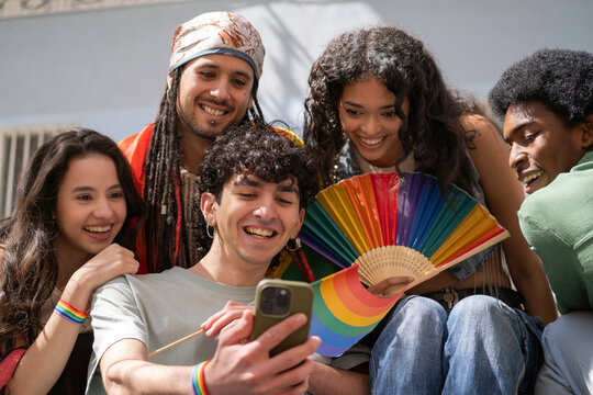 Multiethnic group of cheerful multi-ethnic young people showing lgbtq pride flag and rainbow fan, using smartphone outdoors