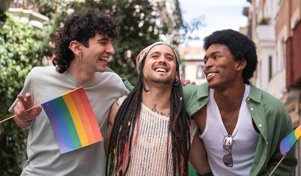 Three multi-ethnic male friends are laughing and walking together during a gay pride parade, holding rainbow flags