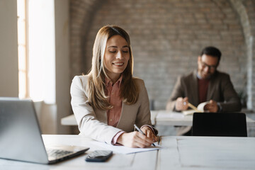 Businesswoman smiling while writing notes during a meeting. She is sitting a desk with laptop,...