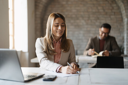 Businesswoman smiling while writing notes during a meeting. She is sitting a desk with laptop, while a colleague in the background is also taking notes in a modern office