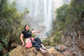 Smiling Asian woman sitting on rock with waterfall in background at Indonesia rainforest. Travel...