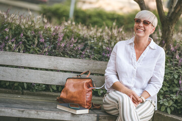 Middle aged woman with blond short hair in sunglasses sitting on bench. Cheerful female person looking to side dressed in white summer shirt while sitting on bench. Outdoors.