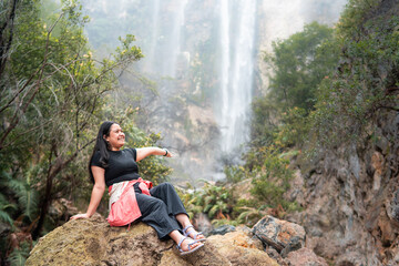 Smiling Asian woman sitting on rock with waterfall in background at Indonesia rainforest. Travel...