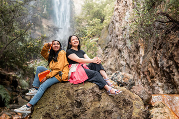 Naklejka premium Two Indonesian women sitting on a large rock, smiling and enjoying friendship in front of a tropical waterfall in Indonesia. Perfect for themes of travel, friendship, adventure, and nature lifestyle.