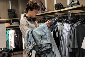 Teen boy 16 years old shopping in apparel store, looking at jeans and denim clothes, modern teenager fashion lifestyle. Adolescent boy in clothing shop choosing casual wear, youth style.