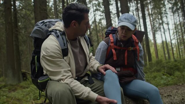 Handheld shot of interethnic man examining sprained leg of his girlfriend sitting on fallen tree during hiking trip in forest