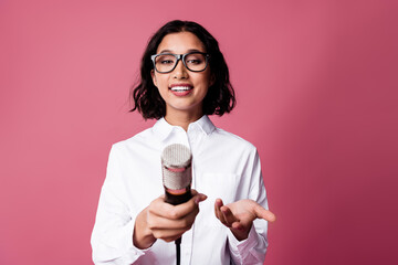 Confident young woman holding a microphone against a pink background, showcasing a stylish and...