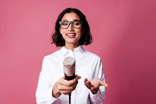Confident young woman holding a microphone against a pink background, showcasing a stylish and professional demeanor in formal attire