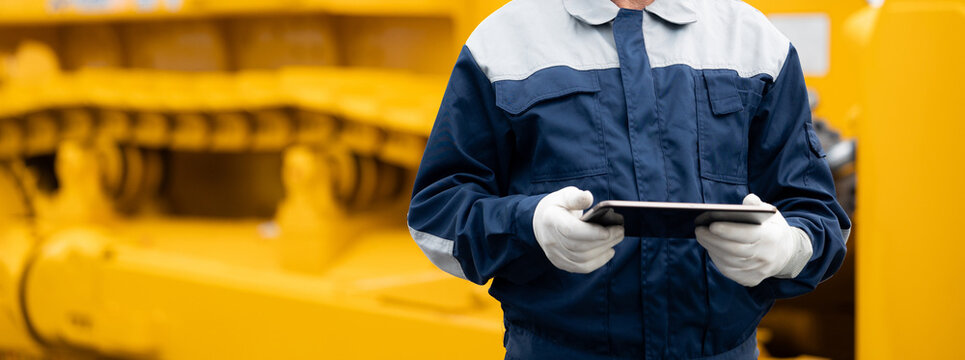 Industrial worker male senior in helmet holds tablet computer on background of production of excavator factory. - Powered by Adobe