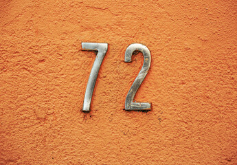 Metallic house number 72 fixed on a textured orange wall, showing weathered details and urban...