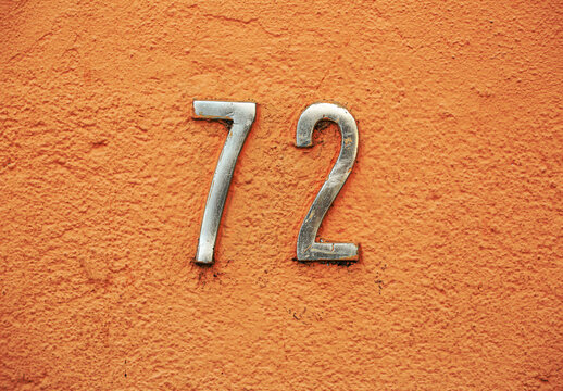 Metallic house number 72 fixed on a textured orange wall, showing weathered details and urban character. A bold numeral composition perfect for design, signage, architecture, or street photography