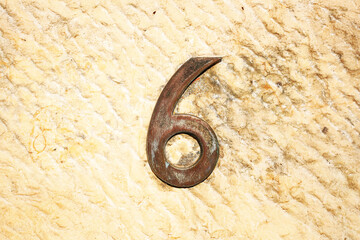 Rustic metallic number 6 mounted on a rough, yellowish stone wall. The numeral shows signs of aging...