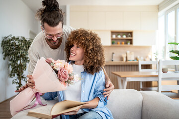 Man giving bouquet of flowers to smiling woman reading book on sofa