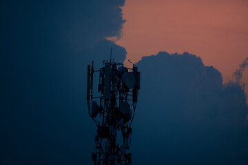 Silhouetted cell tower and antennas against a sunset sky with dark clouds.