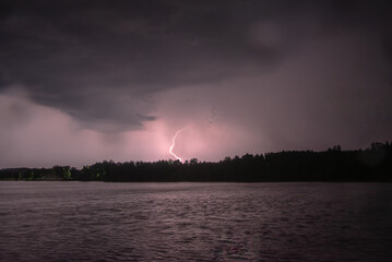 \Lightning strikes over a dark sky above a lake, illuminating clouds and water with a purple glow.
