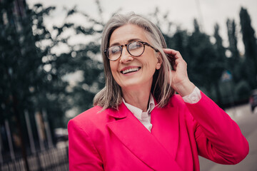 Stylish senior woman outdoors wearing a bright pink suit and smiling confidently in an urban city...