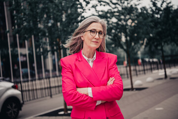 Confident mature woman in a vibrant pink suit posing outdoors on a city street, exuding elegance...