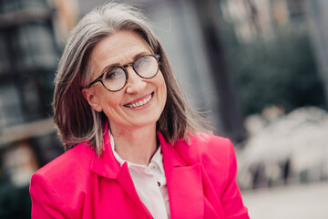 Confident senior woman with gray hair in stylish pink blazer smiling outside in a city setting