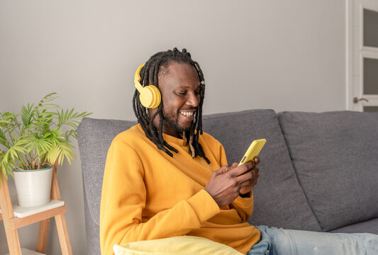 African american man with dreadlocks and yellow headphones using smartphone while sitting on a sofa