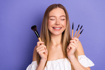 Young woman smiling while holding makeup brushes on a purple background, expressing happiness and...