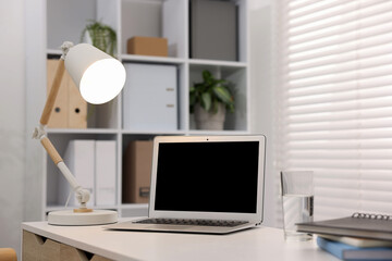 Stylish workplace with laptop, lamp and glass of water on white table in office