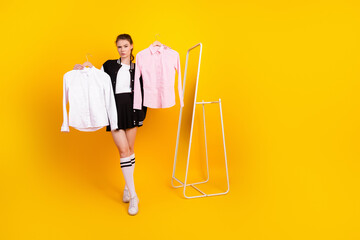 Charming woman in stylish casual wear holding shirts on hangers standing against a bright yellow...