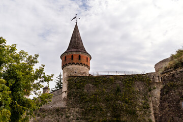 Fototapeta premium Medieval Kamianets-Podilskyi fortress with stone walls and a distinctive tower surrounded by lush greenery and dramatic clouds, showcasing historical architecture and natural beauty